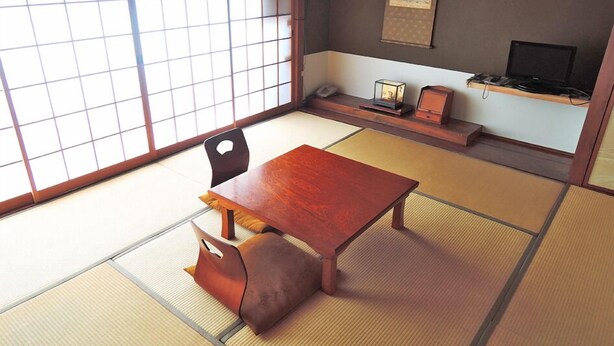 A traditional Japanese room with tatami flooring, a low wooden table, a cushioned bench, a black chair, a small shelf with a lamp, and a television on a stand. - Yayoi Ryokan (Ono)