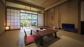 Ein traditioneller japanischer Raum mit Tatami-Boden, einem niedrigen Tisch mit Kissen, einem Fernseher und einer Schiebetür mit Blick auf Grünflächen.