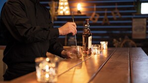 A person pouring a drink into a glass on a wooden bar with candles and bottles in the background. - Etowa Kasama (Kasama)