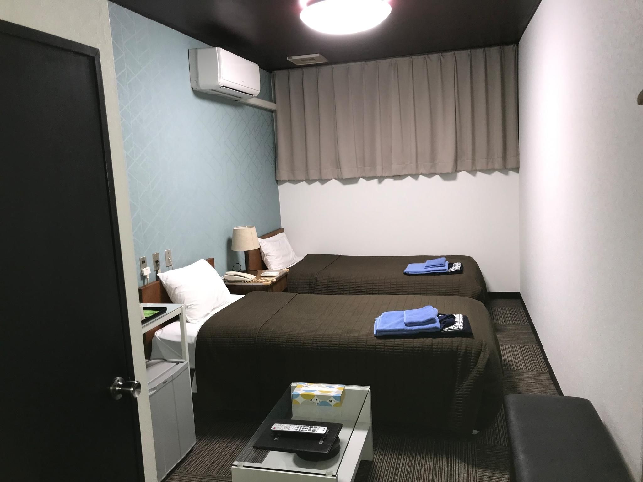 A hotel room with two beds, a desk, a chair, a TV, and an air conditioning unit.