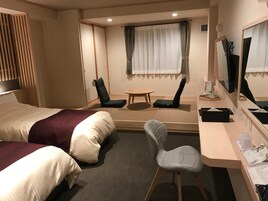 Habitación de hotel con dos camas, un escritorio, una silla, una mesita, un espejo y una ventana con cortinas.