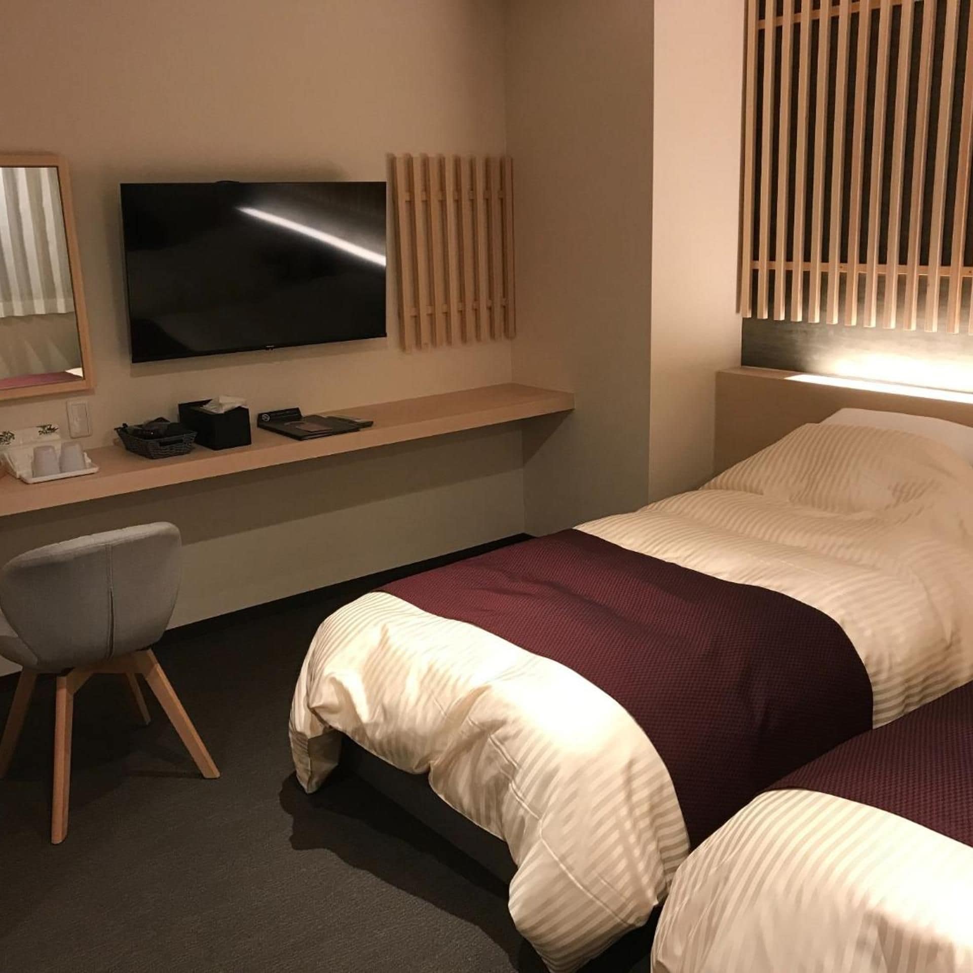 A hotel room with a bed, a desk with a flat-screen TV, a chair, and a window with blinds.