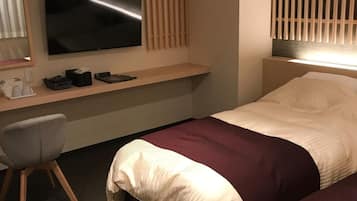 A hotel room with a bed, a desk with a flat-screen TV, a chair, and a window with blinds.