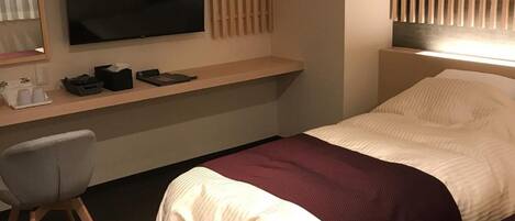 A hotel room with a bed, a desk with a flat-screen TV, a chair, and a window with blinds.