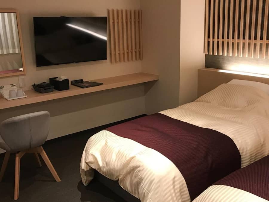 Quarto de hotel com cama, escrivaninha, televisão de tela plana, cadeira e janela com persianas.