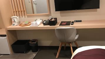 Quarto de hotel com escrivaninha, televisão, cadeira, cama e um pequeno cesto de lixo.