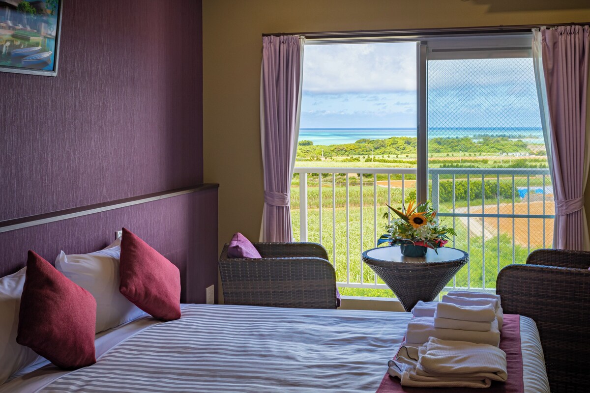 A hotel room with a bed, two chairs, a table with a vase, and a view of the ocean and greenery.