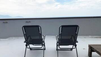 Two black lounge chairs on a rooftop terrace with a view of the sea and mountains.