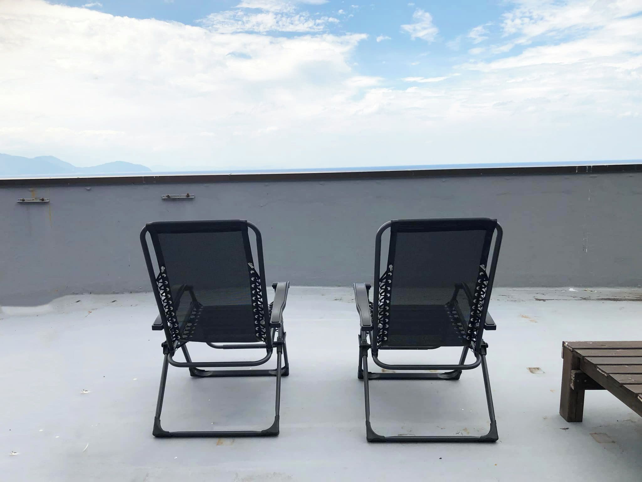 Two black lounge chairs on a rooftop terrace with a view of the sea and mountains.