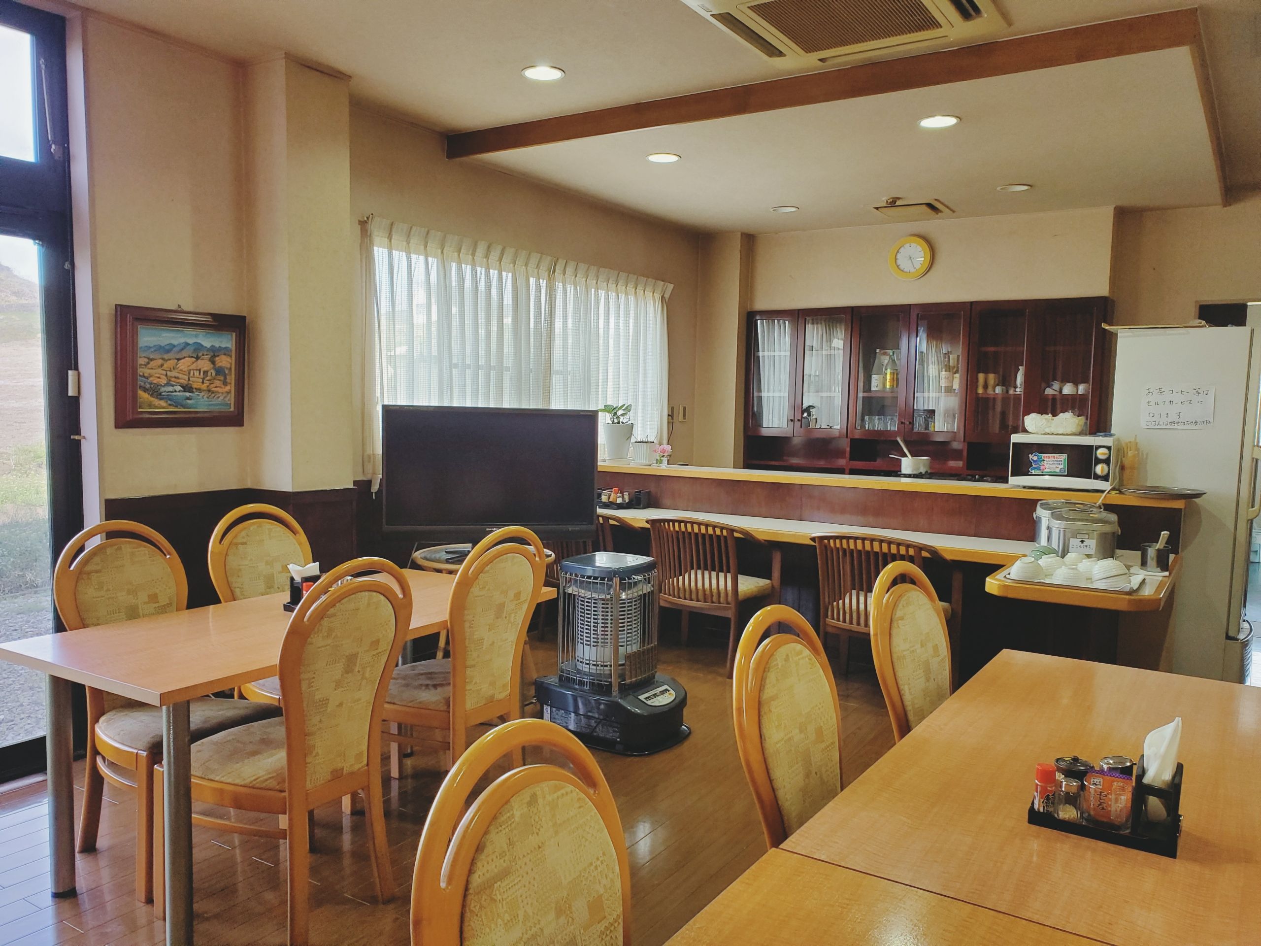 Comedor con mesas y sillas de madera, televisor, zona de cocina con armarios y un reloj en la pared.