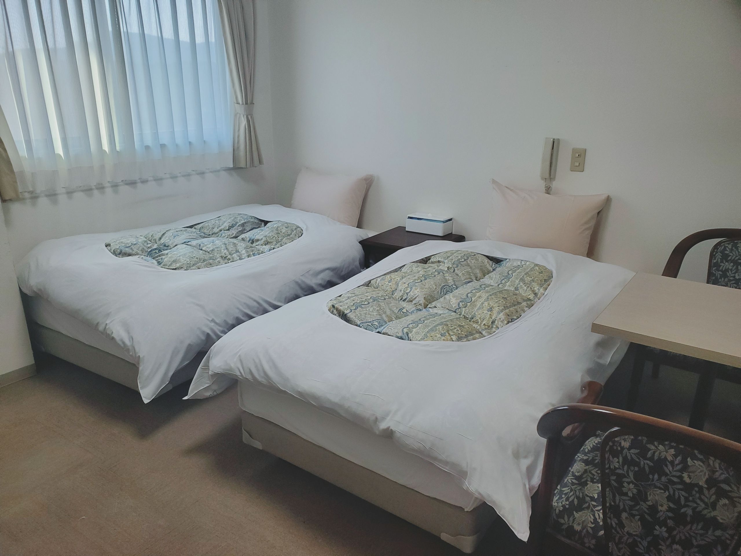 A twin room with two beds, a small table, a chair, and a window with curtains.