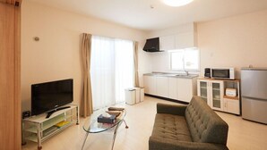 A living room with a flat-screen TV, a glass coffee table, a sofa, a kitchen area with cabinets and appliances, and a window with curtains. - Rainbow Terrace (Ginowan)