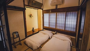 Une pièce de style japonais traditionnel, avec un sol recouvert de tatamis, deux lits simples, une petite table ornée d’un vase et une fenêtre à panneaux coulissants de type shoji.