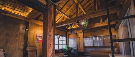 A rustic cabin interior with wooden beams, a window with a view, a bench, and a hanging plant.