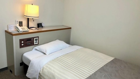 A hotel room with a single bed, a bedside table with a lamp, a phone, and a small control panel.