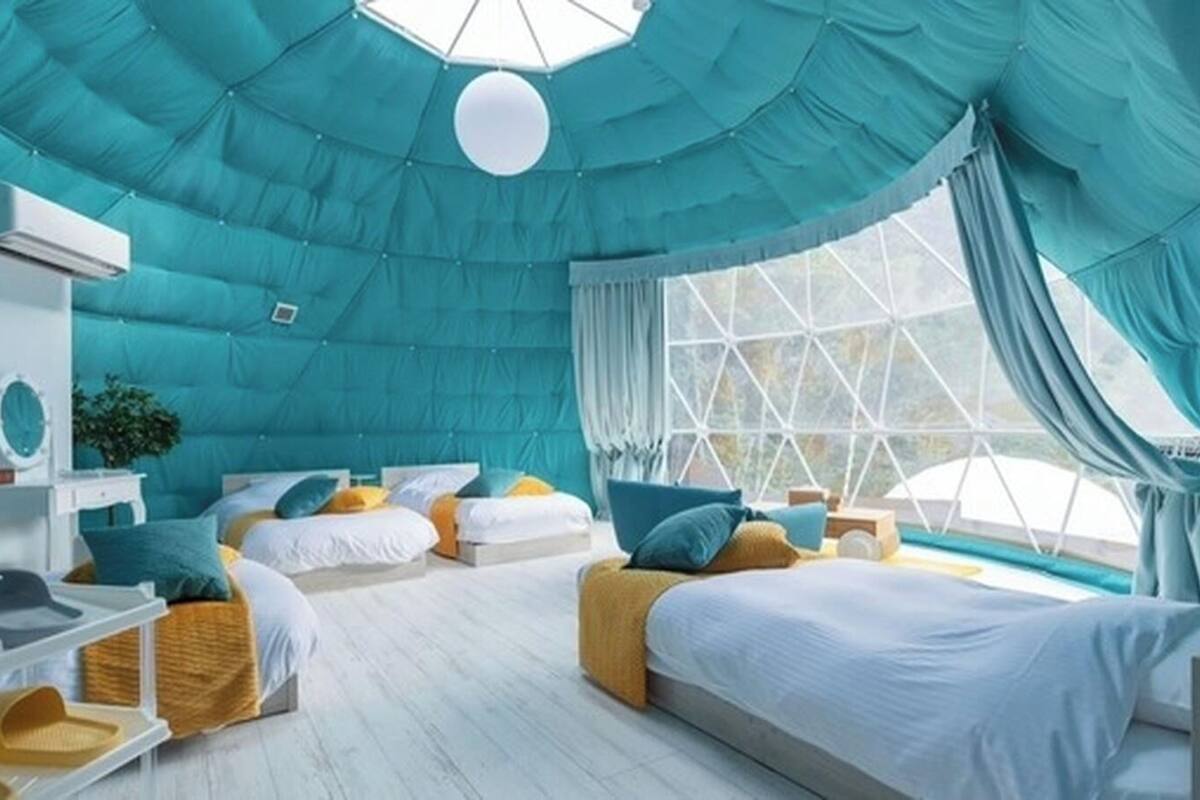 Una habitación con techo abovedado, varias camas, un tocador blanco y un ventanal con cortinas.