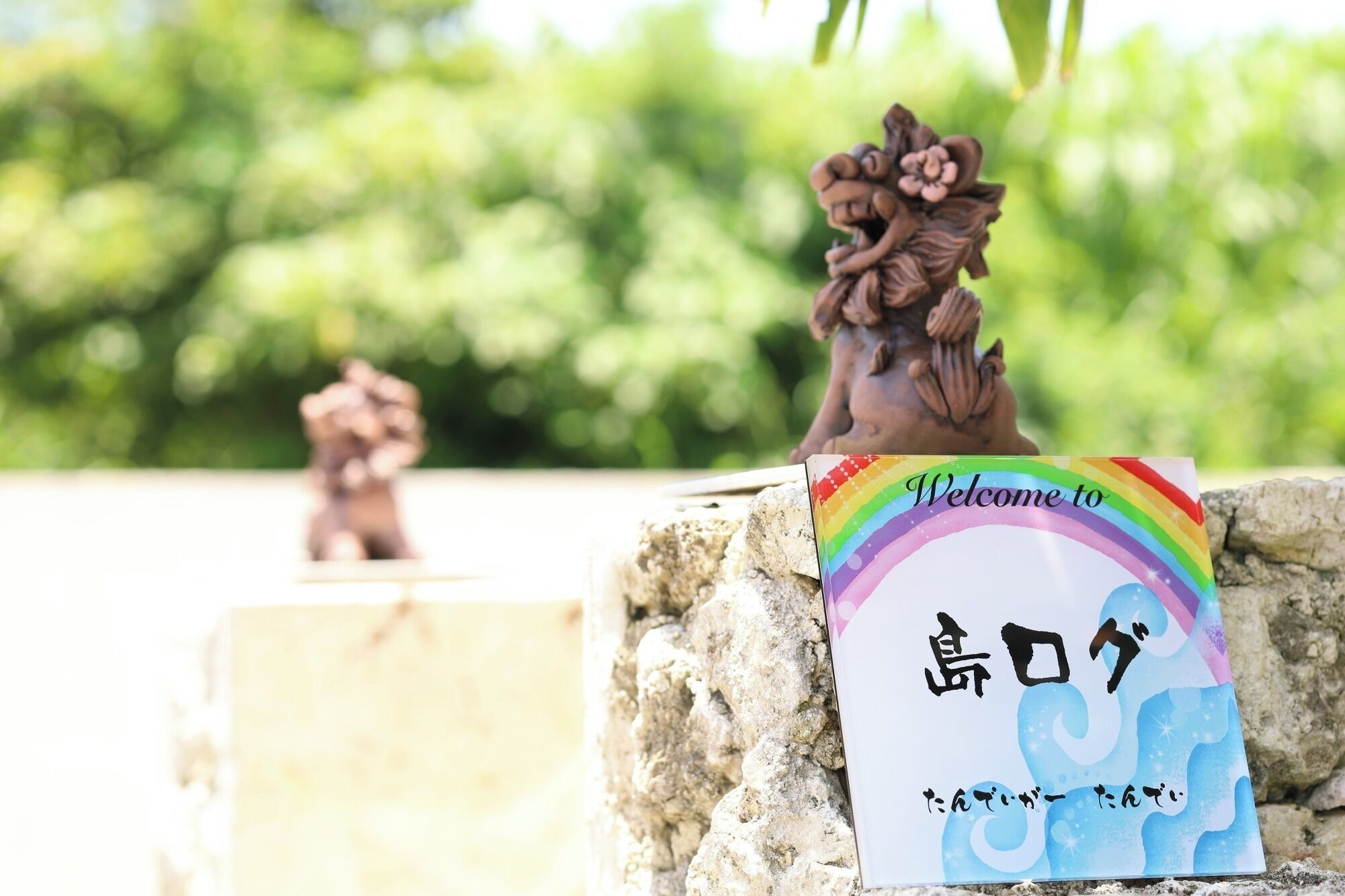 A welcome sign with a rainbow and Japanese characters, placed on a stone structure with a blurred green background.