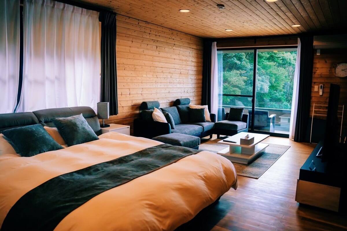 Une chambre d’hôtel moderne, dotée d’un grand lit, de deux canapés, d’une table basse et offrant une vue sur la verdure par la fenêtre.