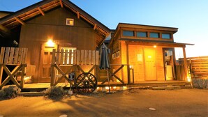 A wooden cabin with a porch, a wheelbarrow, and a sign indicating it is a cabin rental. - Tomareru Himitsukichi L-base (Matsumoto)