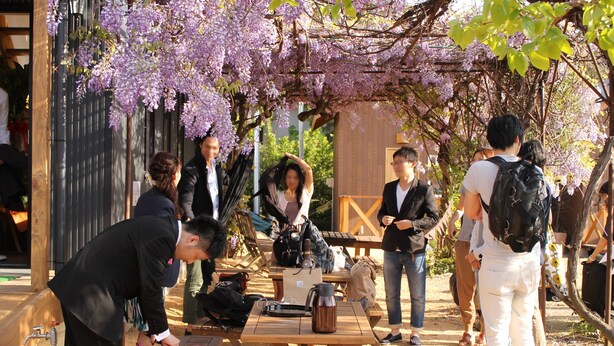 A group of people gathered around a wooden table with a kettle and a pink bag, under a canopy of purple flowers. - Tomareru Himitsukichi L-base (Matsumoto)