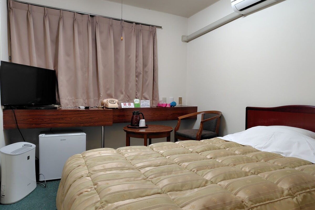 A hotel room with a bed, a desk with a computer, a chair, a small fridge, and a television.