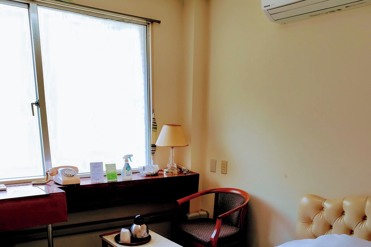 A hotel room with a bed, a desk, a chair, a window, and an air conditioning unit.