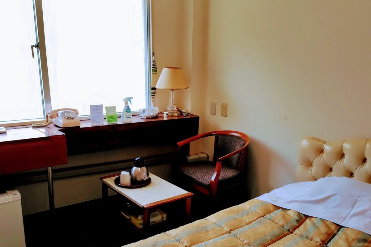 A hotel room with a bed, a desk with a telephone, a chair, a lamp, and an air conditioner.