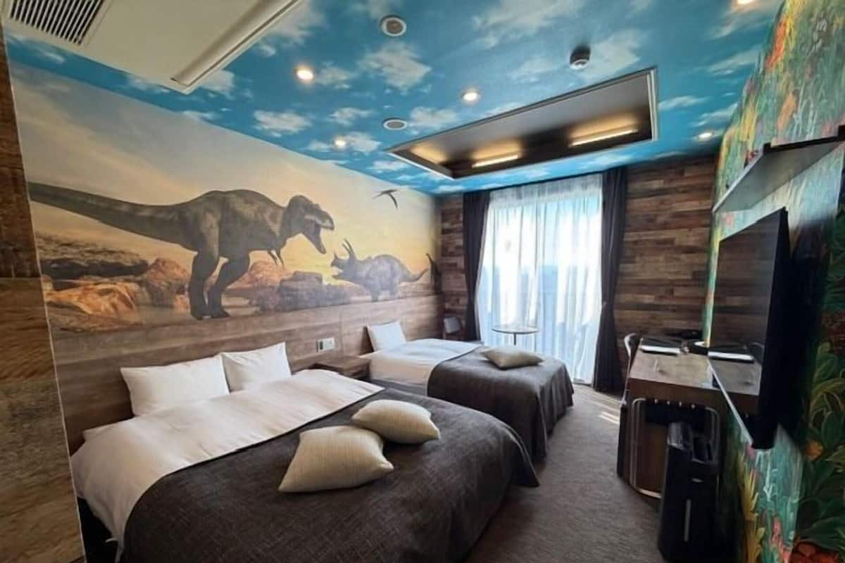 Una habitación con dos camas, una mesita, un televisor y un mural de dinosaurios.