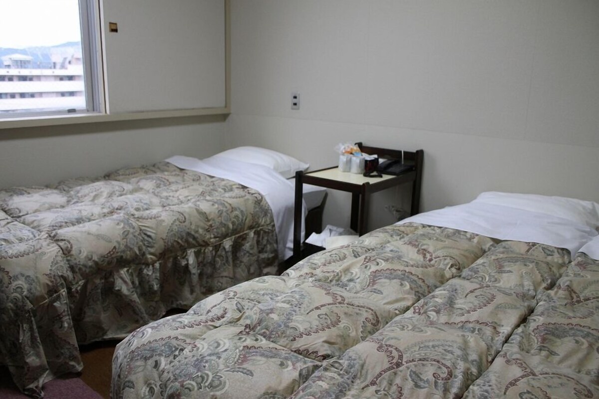 Een kamer met twee bedden, elk voorzien van beddengoed met een patroon, een tafeltje met toiletartikelen, en een raam met uitzicht op gebouwen.