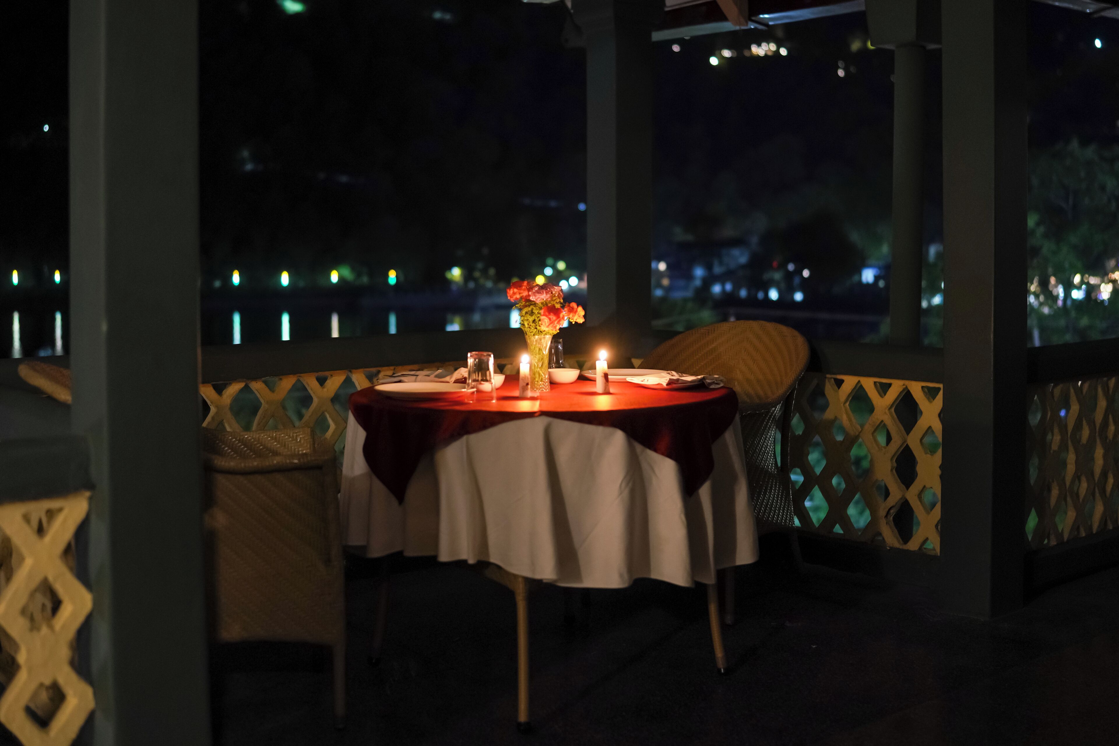 Une table dressée pour deux personnes, avec des bougies, un vase de fleurs et une vue sur le paysage urbain illuminé la nuit.
