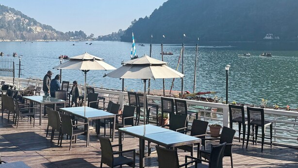 An outdoor dining area with tables and chairs, overlooking a lake with boats and a distant mountain. - The Grand Hotel Nainital (Nainital)