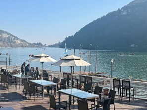 Zona pranzo all'aperto con tavoli e sedie, con vista su un lago con barche e una montagna in lontananza.