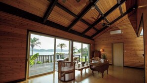 Um quarto espaçoso com paredes de madeira e uma grande porta de vidro deslizante que dá para uma varanda com vista para o mar e palmeiras.