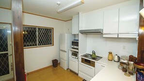 A compact kitchen with white cabinets, a stove, a refrigerator, and a window with a metal screen. - Villa Adanrose Amami (Amami)