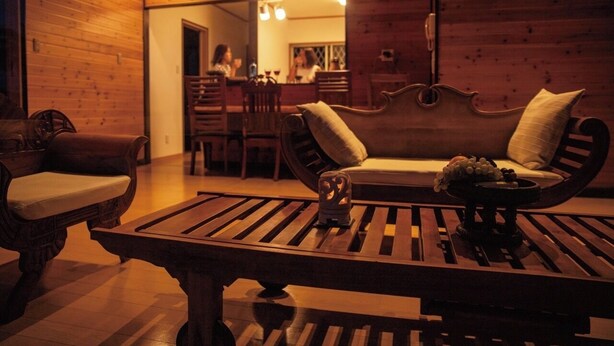 Um interior de madeira acolhedor, com um sofá, cadeiras, uma mesa com uma vela e uma cozinha visível ao fundo.