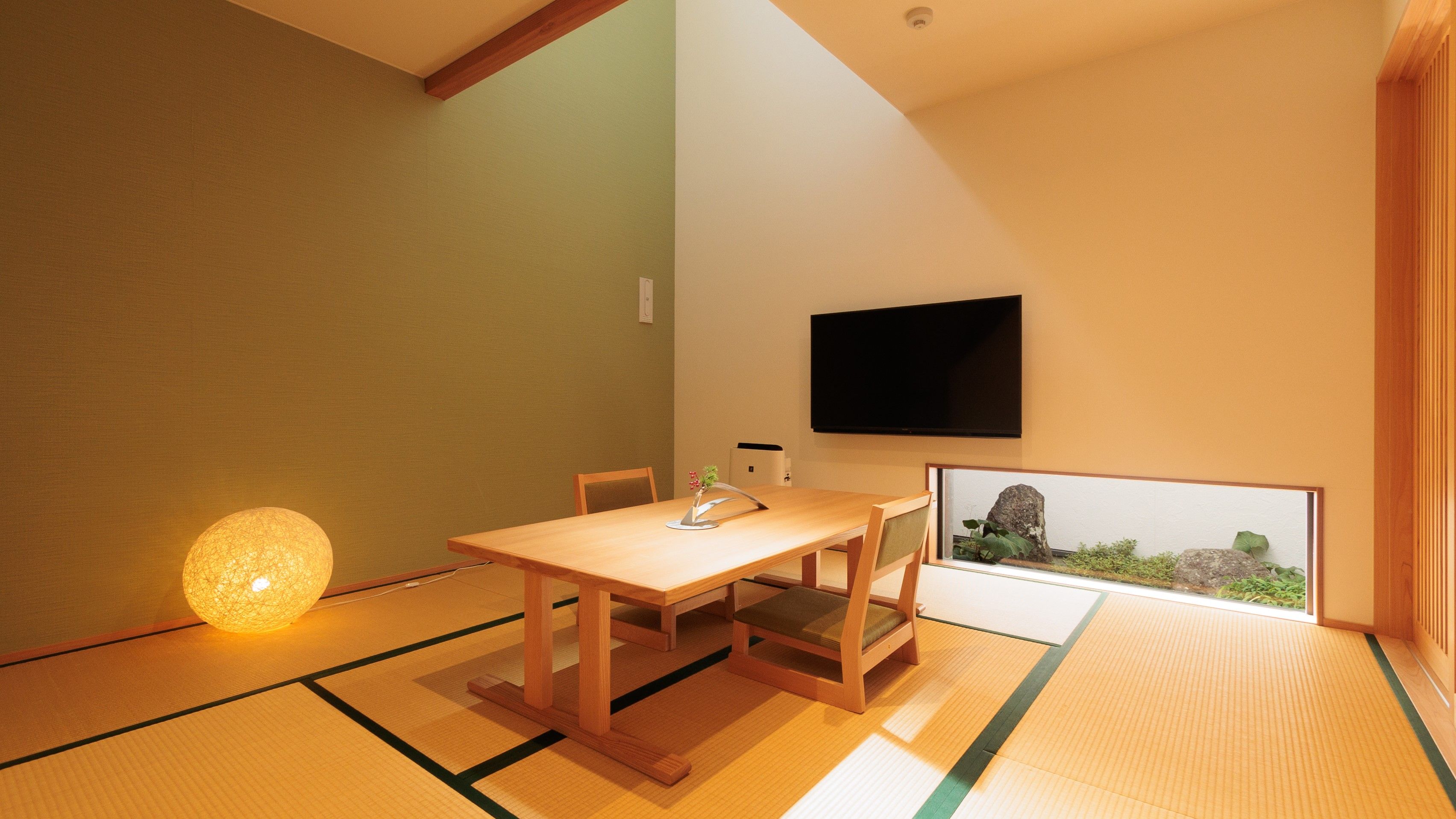 Una habitación japonesa tradicional con suelo de tatami, una mesa baja con cojines, un televisor montado en la pared y un cuadro con un jardín rocoso.