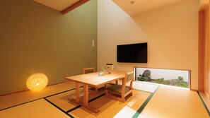 A traditional Japanese room with tatami flooring, a low table with cushions, a television mounted on the wall, and a framed picture of a rock garden.