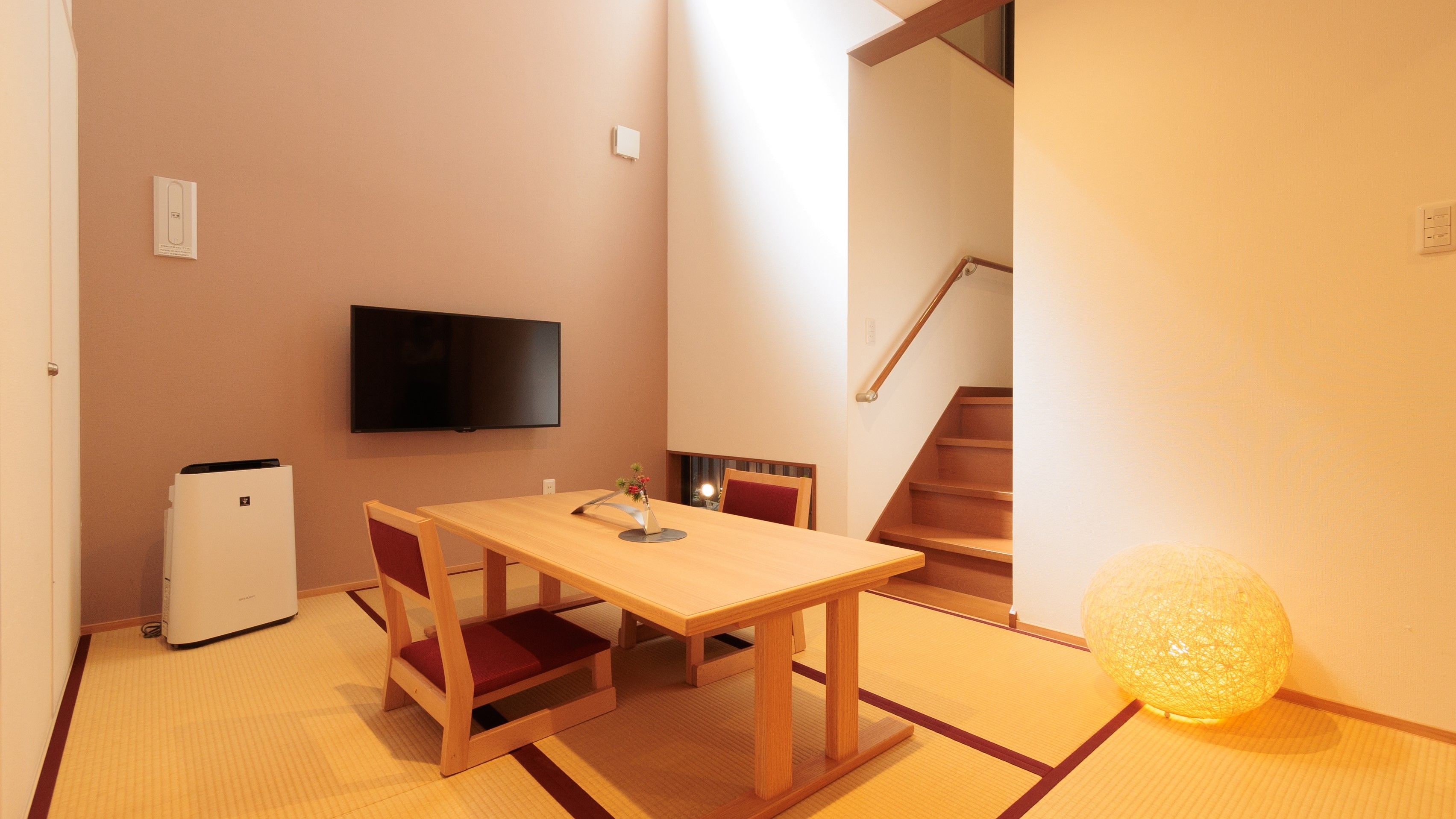 Ein Zimmer mit Holztisch, Stühlen, einem an der Wand montierten Fernseher, einem kleinen Heizgerät und einer Treppe.