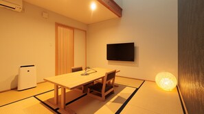A room with tatami flooring, a wooden table, two chairs, a television mounted on the wall, and a small air conditioning unit.