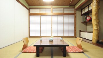 Una habitación japonesa tradicional con suelo de tatami, una mesa baja con cojines, una puerta corredera shoji y una estantería con objetos decorativos.