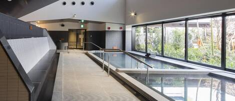 A modern indoor swimming pool with a glass barrier, a staircase on the left, and a view of greenery outside.