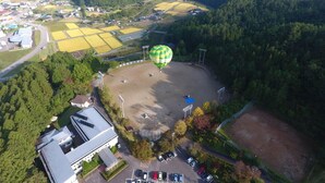 Een luchtballon boven een landelijk landschap met velden, een parkeerplaats en een gebouw met een schuin dak.