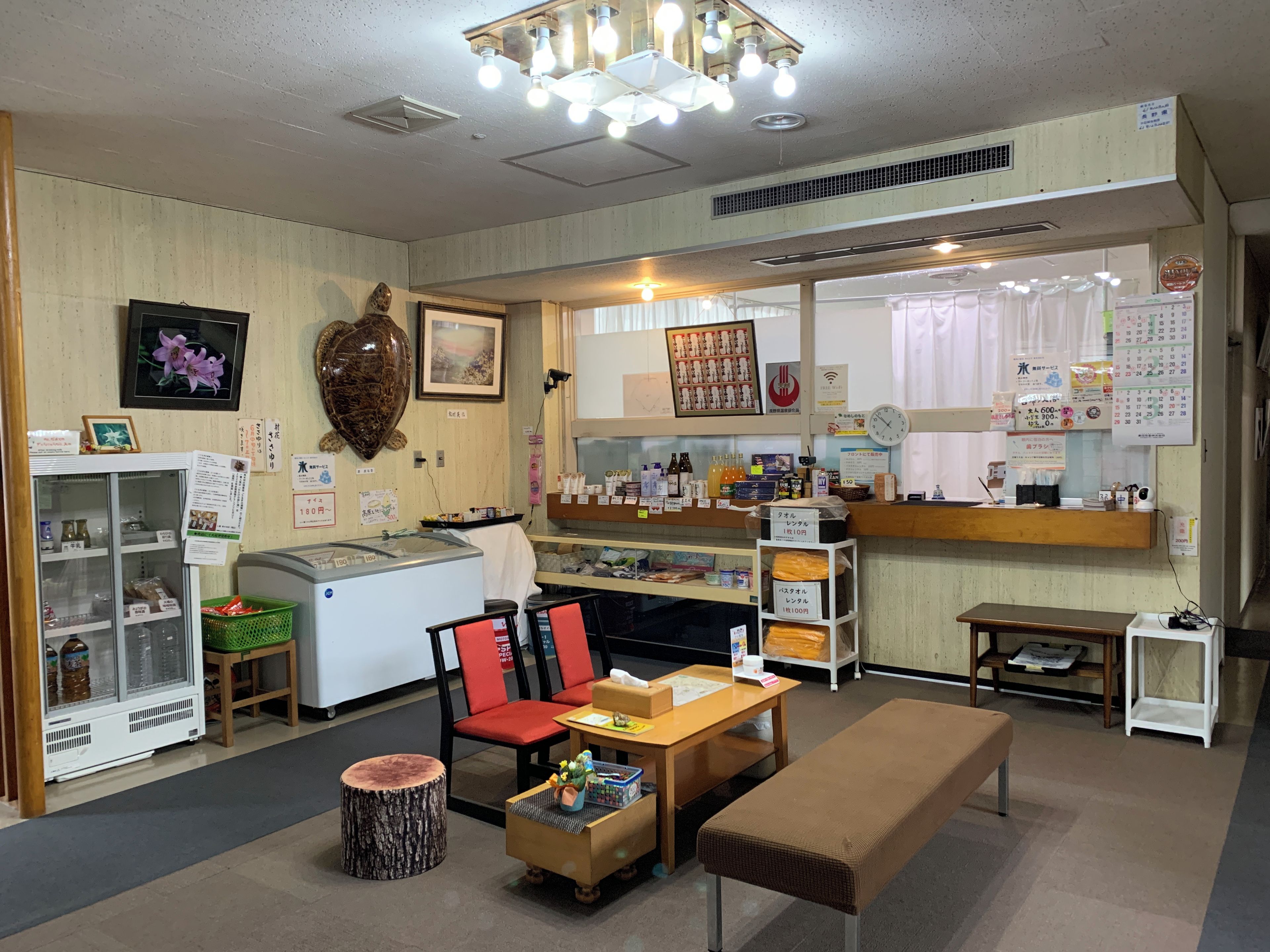 Ein Zimmer mit einer Rezeption, einem Kühlschrank, einer Holzbank und einem kleinen Tisch mit Stühlen.