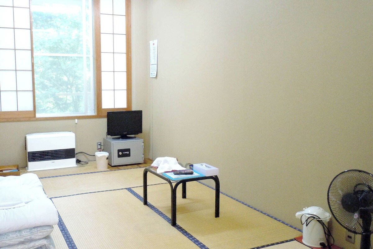 A room with tatami flooring, a bed, a desk with a computer, a television, and a window with a view of trees.