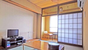 Una habitación japonesa tradicional con suelo de tatami, un televisor sobre un mueble bajo, una puerta corredera de tipo shoji y una pequeña mesa con un objeto redondo encima.
