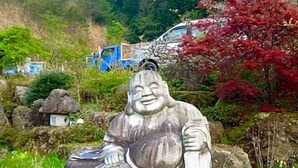 A stone statue of a seated figure in a garden setting with rocks, greenery, and a vehicle in the background.