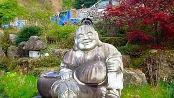 A stone statue of a seated figure in a garden setting with rocks, greenery, and a vehicle in the background.