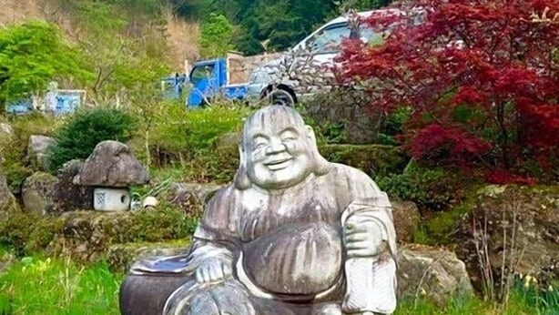 A stone statue of a seated figure in a garden setting with rocks, greenery, and a vehicle in the background. - Minshuku Yurika (Urugi)
