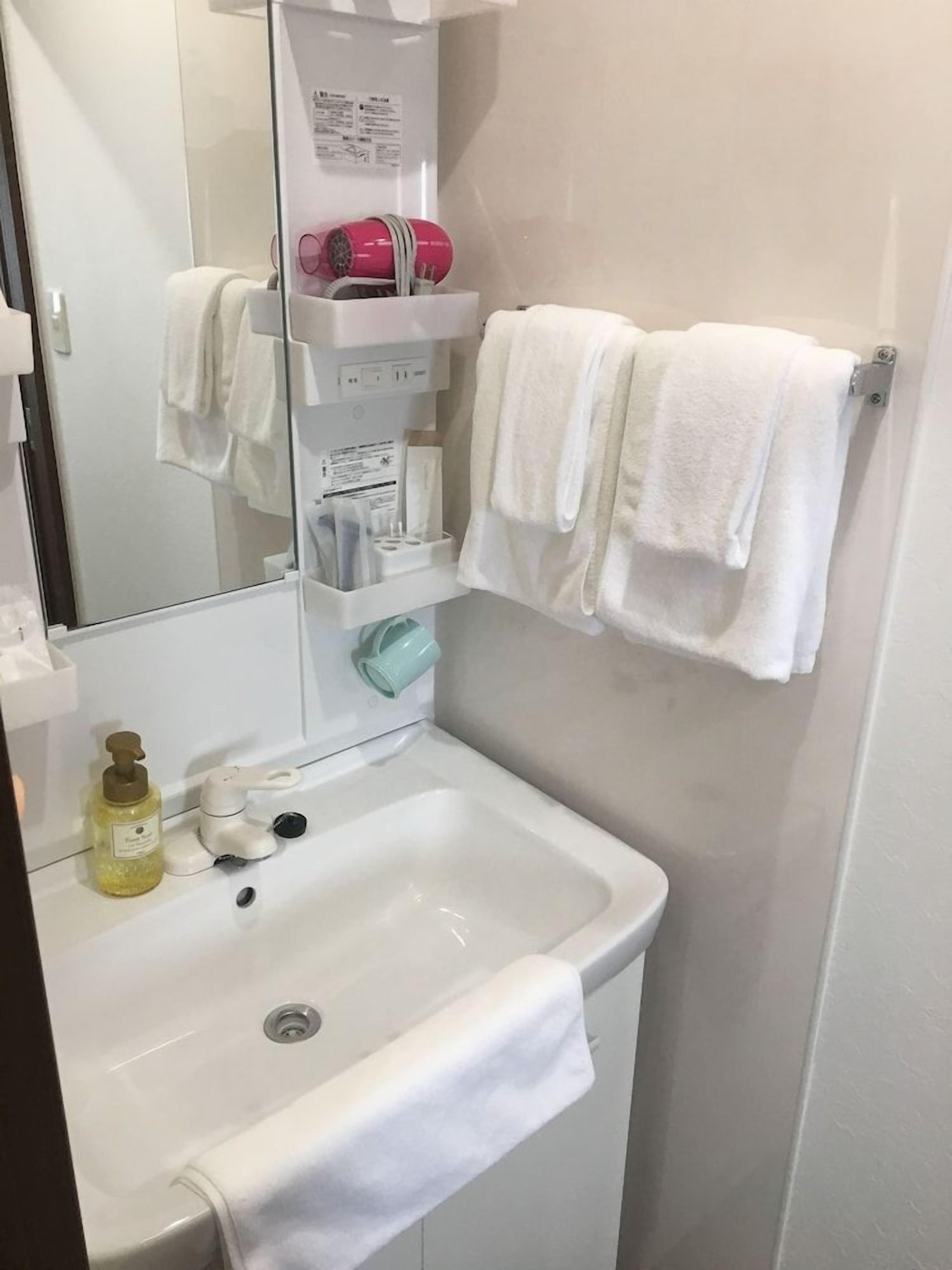 A bathroom with a white sink, a mirror, a towel rack with two white towels, a shelf with personal care items, and a bottle of liquid soap.