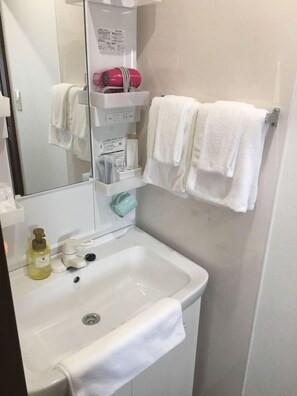 Une salle de bain avec un lavabo blanc, un miroir, un porte-serviettes avec deux serviettes blanches, une étagère contenant des articles de toilette et un flacon de savon liquide.